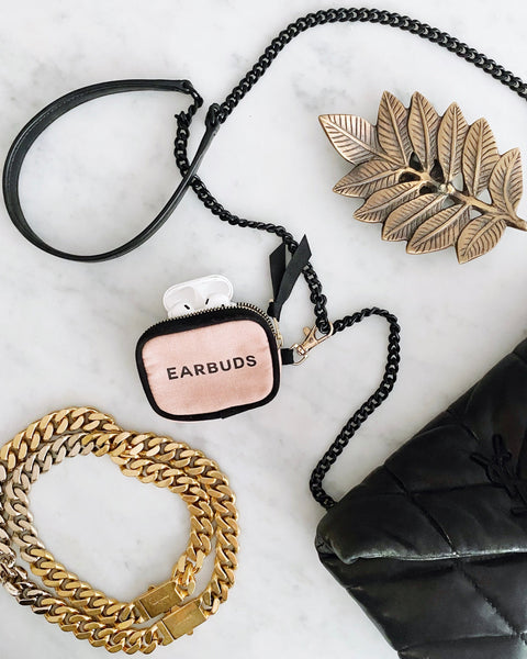 Bag-all Earbuds Case in blush pink with gold clasp and black chain strap, featuring zipper closure and black lettering. Shown with stylish accessories on marble surface