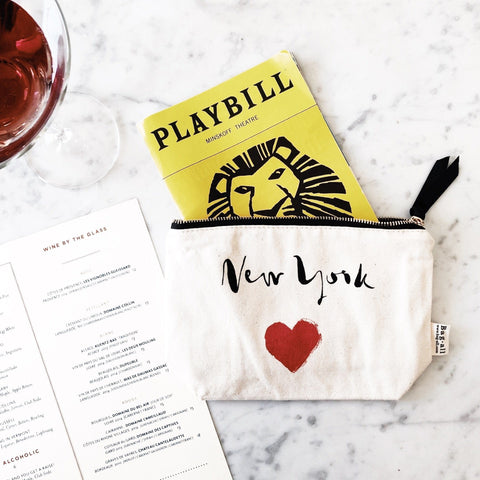 Bag-all New York Heart Pouch in cream canvas with red heart print, black zipper, and Playbill peeking out, displayed on marble surface with wine glass and menu