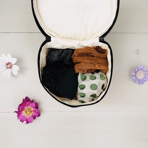 Bag-all Round Lingerie Case in cream, open to display neatly organized undergarments in black, brown, and polka dot patterns, styled with delicate flowers on white wooden surface