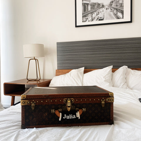 Bag-all Personalized Luggage Handle Cover featuring vintage-style monogram "Julia" on brown leather suitcase, displayed on white hotel bed with modern decor and bedside lamp