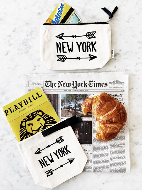 Bag-all New York Arrow Pouch in cream canvas with black arrow design, shown with Metro card and NYC essentials on marble surface against New York Times newspaper and Playbill