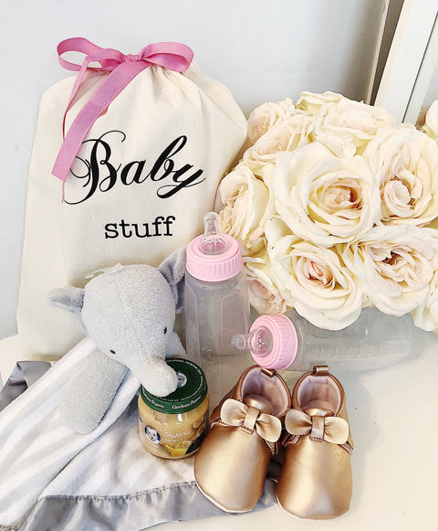 Bag-all Baby Fancy Bag in Cream with pink ribbon, surrounded by baby essentials like bottles, plush toy, and gold baby shoes. Perfect for gifting