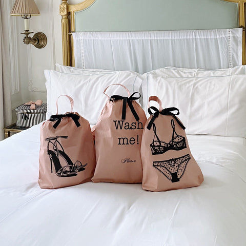 Bag-all Pink Polkadot Lingerie Travel Bags featuring elegant black illustrations of heels, wash me text, and lingerie designs, displayed on white bedding with satin drawstring closures and black bows