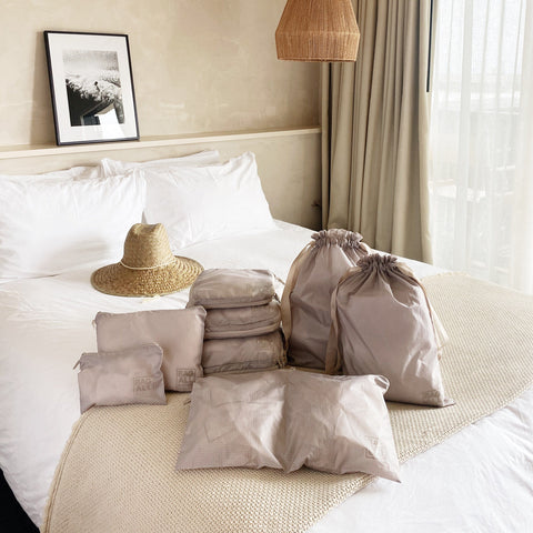 Bag-all Compression Cubes & Packing Bags Set in taupe, featuring 8 eco-friendly nylon organizers displayed on white bedding with straw hat, perfect for sustainable travel