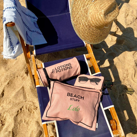 Bag-all Stylish Beach Essentials 3-pack in pink blush featuring monogrammable waterproof pouches for beach items, sunglasses, and lotions, displayed on blue beach chair with straw hat in sunlit sand