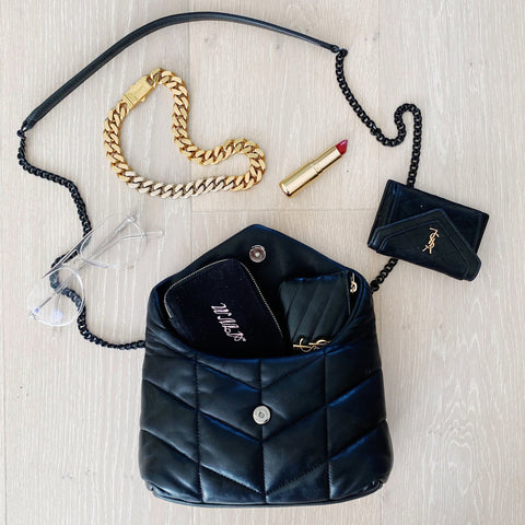 Bag-all Jewelry/Trinket Box in black with gold zipper, displayed alongside gold chain bracelet, lipstick and designer accessories on white wooden surface, perfect for organizing small valuables