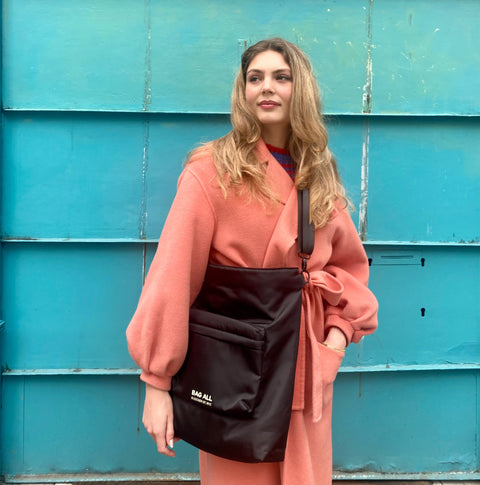 Bag-all City Backpack/Tote in black recycled nylon shown against turquoise wall, featuring convertible straps, padded laptop protection, and spacious front pocket.