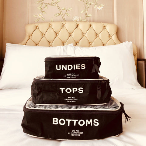 Bag-all Cotton Packing Cubes 3-pack in Black with white mesh and text labels for Undies, Tops, and Bottoms, stacked on white bed against tufted headboard