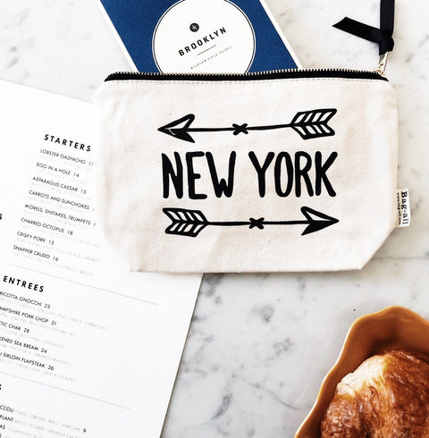 Bag-all New York Arrow Pouch in cream canvas with black arrow design and text, featuring metal zipper, displayed on marble surface next to restaurant menu and pastry