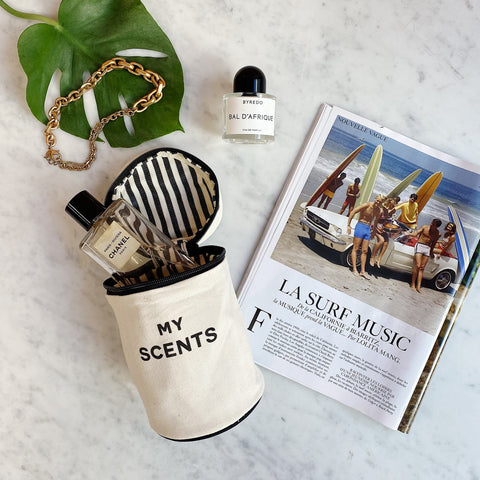 Bag-all Round My Scents Case in cream with black striped interior, stylishly displayed with perfume bottles, gold chain accessory, and monstera leaf on marble surface