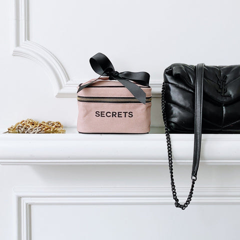 Bag-all Mini Beauty Box for Secrets in blush pink cotton with black bow, double zippers and handle, styled on white shelf next to black handbag
