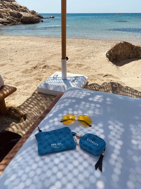 Bag-all Earbuds/Airpods Case with Clasp in Denim blue shown on beach lounger with yellow sunglasses, turquoise ocean and rocky shore in background, demonstrating perfect summer accessory for device protection