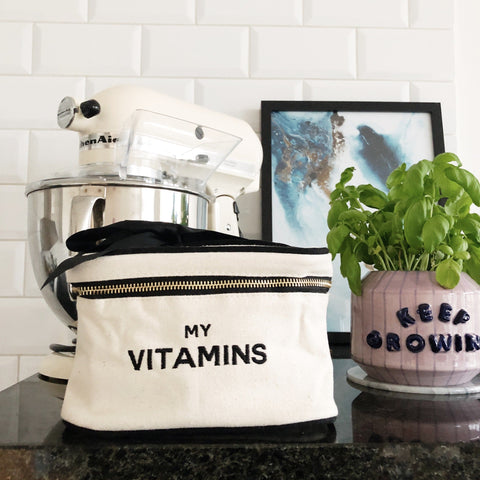 Bag-all Vitamins Storage and Travel Box in cream canvas with black embroidered text, displayed on kitchen counter next to stand mixer and potted basil plant