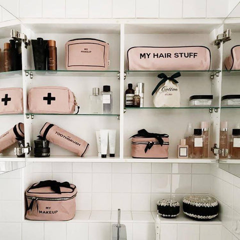 Bag-all My Makeup Pouch in blush pink displayed in organized bathroom shelving with other matching pink beauty cases and toiletries, featuring black text and gold zipper details
