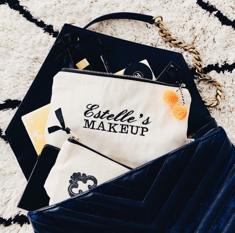 Canvas makeup bag with personalized embroidery reading Estelle's Makeup placed inside a black handbag on a rug