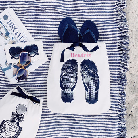 Bag-all Cream Flip Flops Shoe Bag with navy ribbon accent shown on striped beach blanket with sunglasses and travel accessories, perfect for beach organization and summer storage