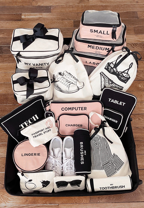 Bag-all black toothbrush travel case displayed with collection of branded organizing bags in pink, black and white featuring labeled compartments for tech, toiletries and clothing essentials on wooden floor