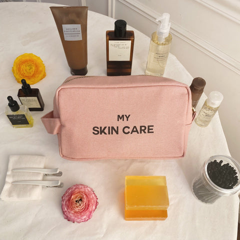 Bag-all My Skin Care Organizing Pouch in blush pink cotton with waterproof lining, surrounded by beauty products, flowers, and skincare essentials on white surface