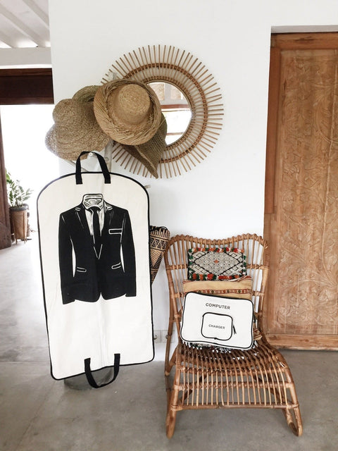 Bag-all Men's Suit Garment Bag with Pocket in cream, featuring black suit design, dual handles, and storage pocket, displayed against white wall with rattan decor