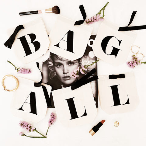 Bag-all Small Letter Bags Classic in cream with black typography letters, arranged with pink flowers, makeup brushes and lipstick on white background, perfect for organizing essentials