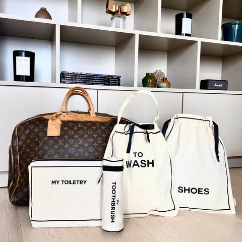 Bag-all Toiletry Travel Gift Set 4-Pack in cream featuring cotton organizing pouches for toiletries, toothbrush, shoes, and washing items displayed next to designer travel bag on white shelf unit