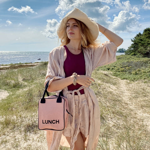 Bag-all pink insulated lunch box with black trim and LUNCH text shown at beach, perfect for stylish meal transport. Professional design with durable metal zipper and padded handle.