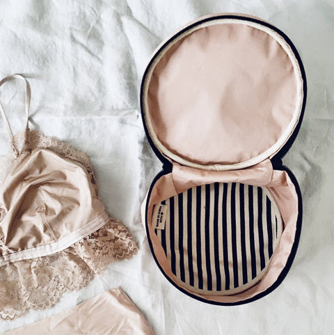 Bag-all Round Lingerie Case in blush pink with black and white striped interior lining, shown open next to delicate lace undergarments on white background