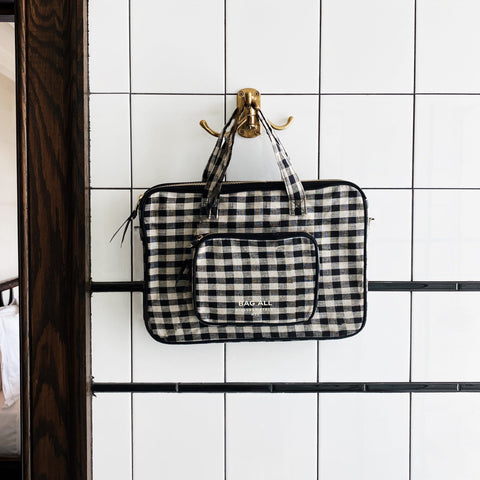 Bag-all Laptop Case 13" in black and white gingham pattern with handle, charger pocket, and protective padding, hanging on white tiled wall, showcasing versatile design