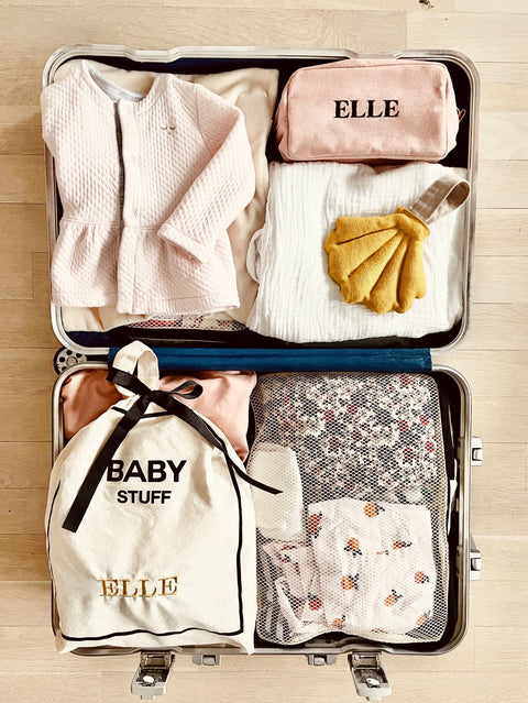 Open suitcase packed for a weekend trip, featuring Bag-all Baby Couture Bag in Cream and makeup pouch, both personalized with ‘ELLE’ monogram