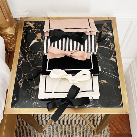 Bag-all Couture Jewelry Roll Medium Black: Elegant travel organizer with decorative bows in pink, black, and white, displayed on marble-top gold table, featuring striped pattern and multiple compartments