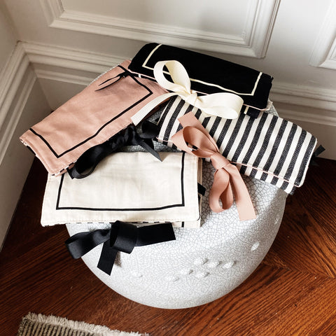 Bag-all Couture Jewelry Roll Medium in pink with black and white striped lining, featuring three zipper compartments, leather earring holder, and padded ring storage on a decorative white ceramic surface