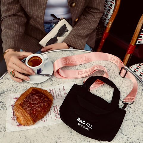 Bag-all Mini Tote Bag with Strap shown in black cotton with pink adjustable strap, displayed on cafe table next to coffee and pastry, highlighting compact size and everyday versatility