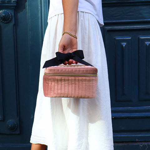 Hand holding a personalized quilted pink velvet makeup cosmetic box with a black bow handle against a dark door background