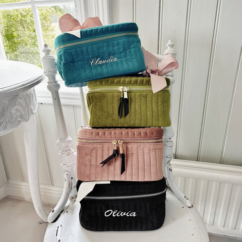 Stack of personalized quilted velvet cosmetic boxes in teal, green, pink, and black on a white chair near a window.
