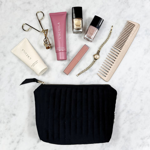 Quilted black velvet makeup pouch with gold zipper, displayed with cosmetics, nail polish, cream, comb, and watch on marble surface