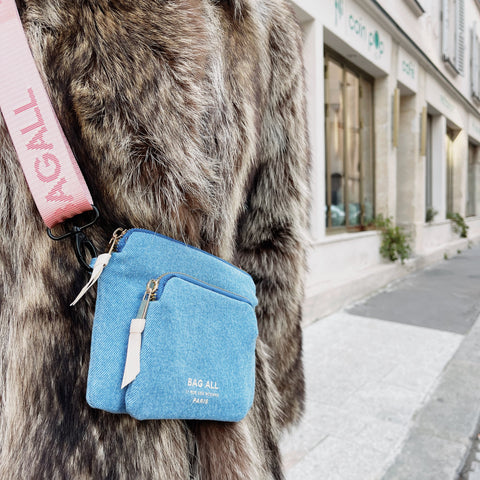 Bag-all Padded Trinket Pouch Double Pocket in denim blue worn crossbody with pink strap, shown against grey fur coat on street, featuring dual zippered compartments and white leather trim