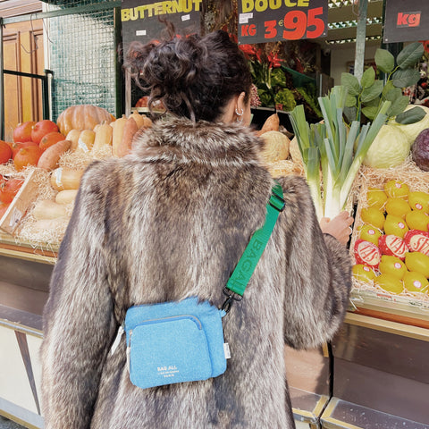 Bag-all Padded Trinket Pouch Double Pocket in Denim worn crossbody with green strap, shown at produce market display with pumpkins and vegetables in background