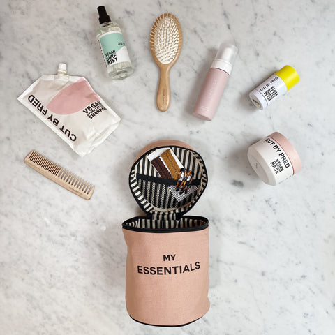 Bag-all Round My Essentials Case in pink/blush shown open with beauty products, featuring striped interior lining, elastic bands, and surrounded by travel-sized toiletries on marble surface