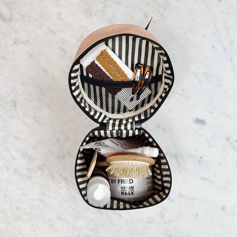 Bag-all Roomy Travel Essentials Case in pink/blush featuring striped interior, organized compartments displaying beauty products, makeup brushes, and skincare items in a round cotton case with waterproof lining