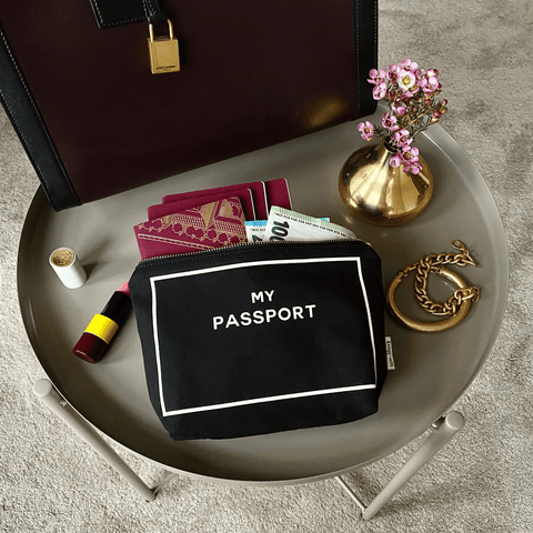 Bag-all black Passport Pouch styled with red passports, cash and travel essentials on grey surface with gold vase and pink flowers, perfect for organized travel