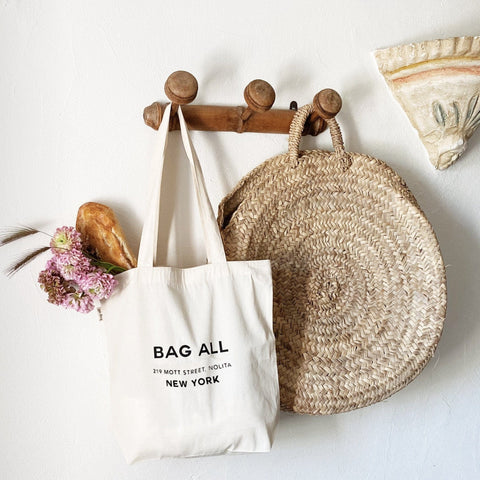 Bag-all New York City Tote hanging on wooden wall hooks alongside decorative straw basket and dried flowers, showcasing minimalist cream cotton design with black text logo