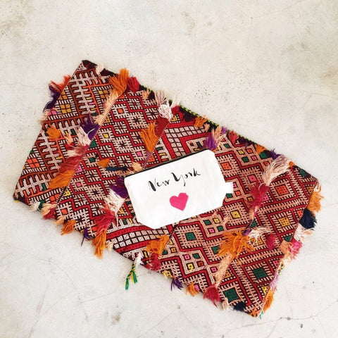 Bag-all New York Heart Pouch in cream displayed on vibrant geometric patterned textile with orange and purple tassels, featuring handprinted design and zip closure