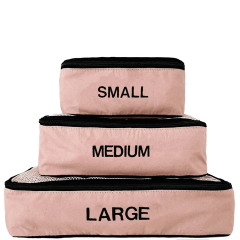 Three pink blush cotton packing cubes in small, medium, and large sizes stacked with black zippers and monogrammed labels.