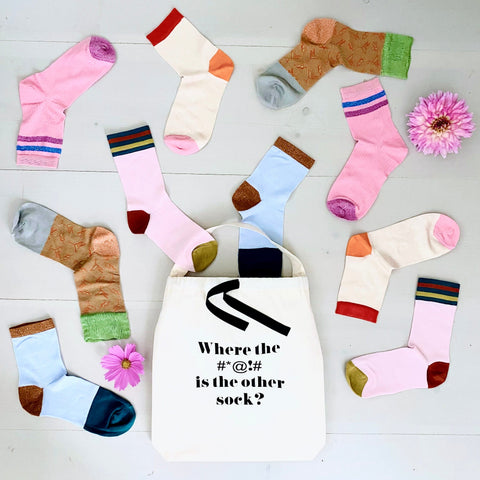 Bag-all Where #*@!# Sock Bag in cream canvas with black text, surrounded by colorful scattered socks and pink flowers on white wooden surface, perfect for sock organization