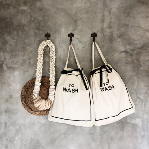 Bag-all To Wash Laundry Bag in cream cotton with black text, featuring drawstring closure and hanging handle, displayed alongside matching bag against textured gray wall