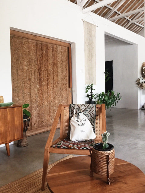 Bag-all Wash Me Laundry Bag in cream color displayed on wooden chair with woven seat, styled in modern minimalist room with carved wooden door and indoor plants