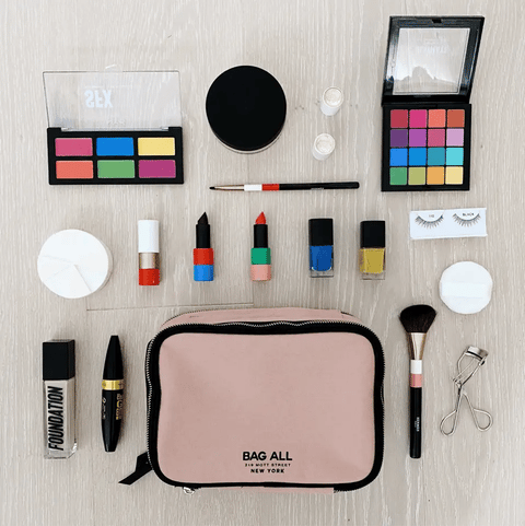 Bag-all Double Sided Toiletry Case in Pink/Blush surrounded by makeup essentials including eyeshadow palettes, lipsticks, nail polishes, brushes, and beauty tools on light wooden surface