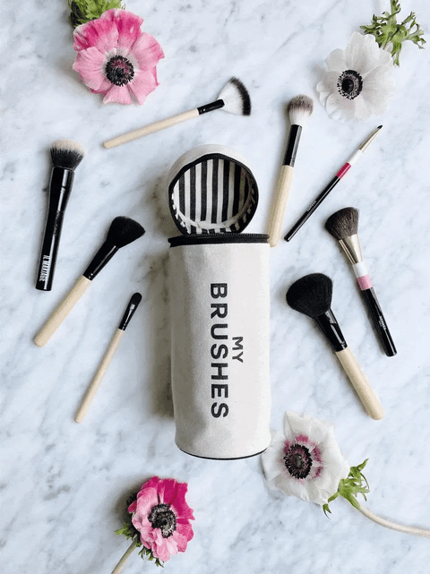 Bag-all Brushes Cylinder Case in cream with striped interior, displayed with makeup brushes and anemone flowers on marble surface, showcasing stylish organization solution