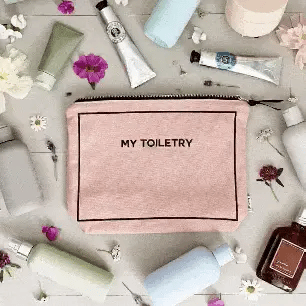 Bag-all Toiletry Pouch with Coated Lining in blush pink, featuring waterproof interior and metal zipper, surrounded by beauty essentials on gray surface with scattered flowers