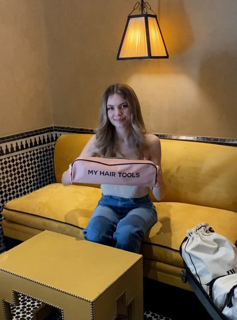 Bag-all Roomy Hair Tools Travel Case in pink blush shown on yellow couch, featuring heat-resistant lining and gold zipper, perfect for Airwrap and styling tools storage